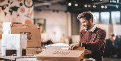 Man zit aan tafel en is een pakket aan het openen. Hij lacht vrolijk en kijkt naar het pakket.