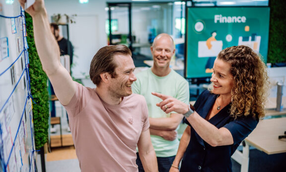 3 collega's staan bij een bord en lachen samen. 1 iemand plakt iets op het bord en iemand wijst naar het bord.